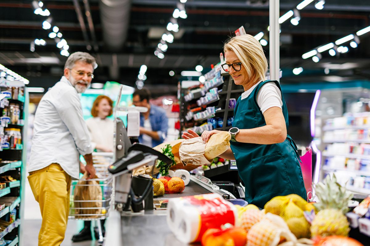Senior female cashier scanning groceries of a customer at checkout counter in a supermarket, with a senior man pushing his shopping cart and waiting to pay for his groceries